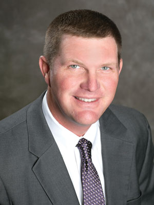 Portrait of Glen Cope MFA Oil board Chairman smiling man with short hair, wearing a gray suit jacket, white shirt, and a patterned tie, set against a blurred dark background.