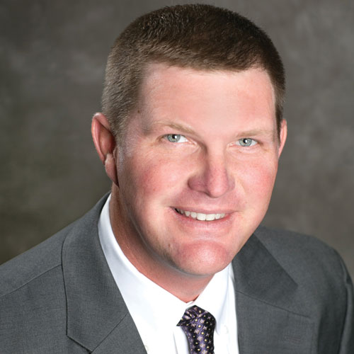 Portrait of Glen Cope MFA Oil board Chairman smiling man with short hair, wearing a gray suit jacket, white shirt, and a patterned tie, set against a blurred dark background.