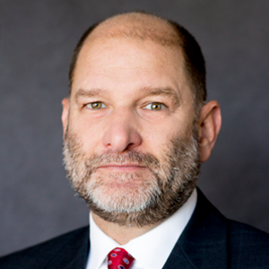 A photo of Tim Danze MFA Oil hedging manager, wearing a dark suit, white shirt, and red tie, against a gray background.