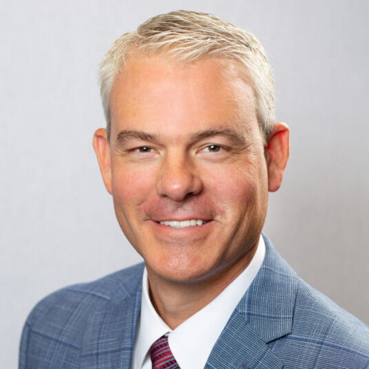 A man with short gray hair in a blue plaid suit jacket, white shirt, and red patterned tie, smiling at the camera against a plain light gray background.