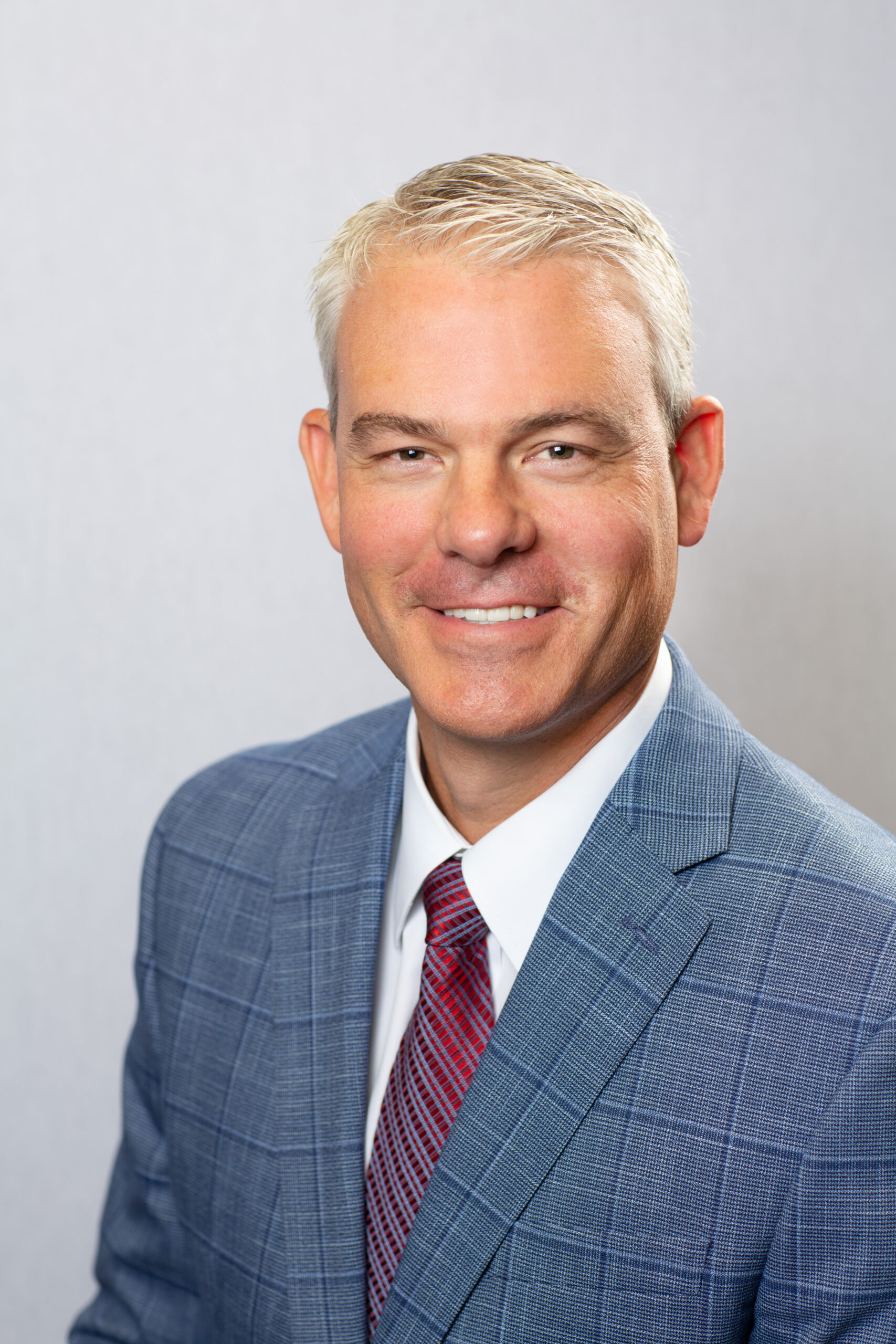 A man with short gray hair wearing a blue plaid suit jacket, white dress shirt, and red patterned tie, smiling at the camera against a light gray background.