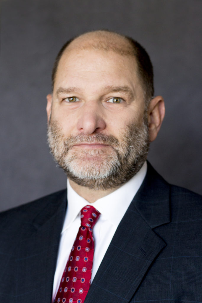 A photo of Tim Danze MFA Oil hedging manager who is wearing a dark suit, white shirt, and red patterned tie. He is standing against a plain, dark background.