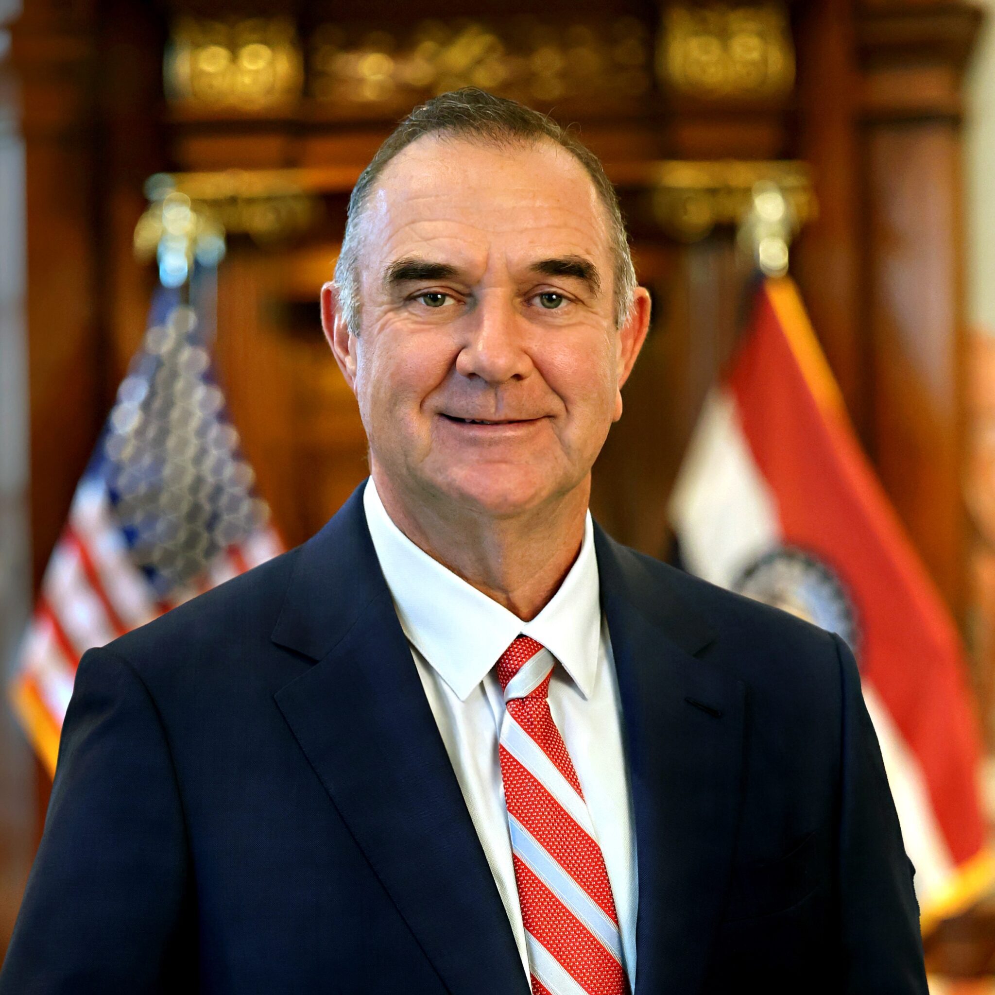A man in a dark suit, white shirt, and red striped tie stands smiling in front of American and Missouri state flags, with a wooden background behind him.