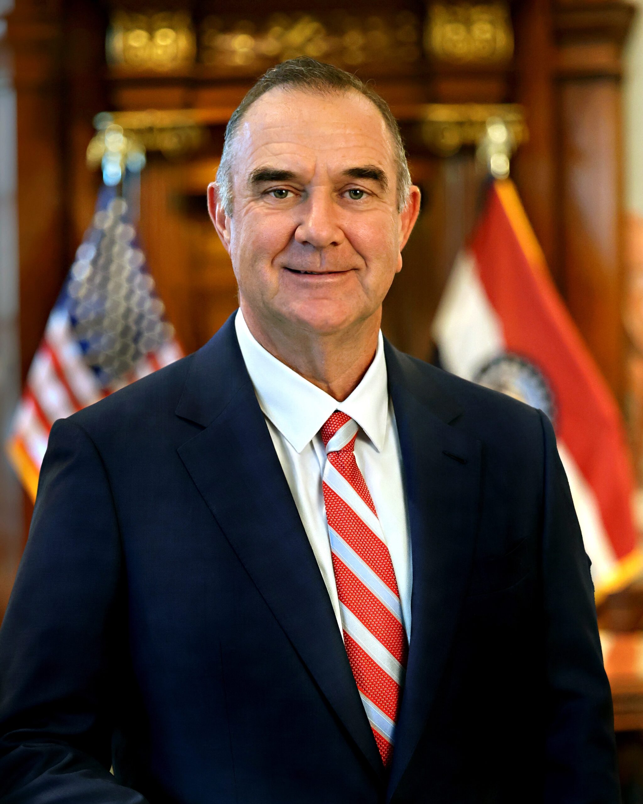A man in a dark suit and red striped tie stands indoors in front of blurred U.S. and Missouri state flags, smiling at the camera.