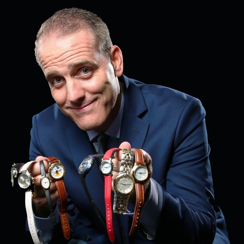 A man in a blue suit smiles while holding out his hands, which are draped with multiple wristwatches of different styles and colors against a black background.