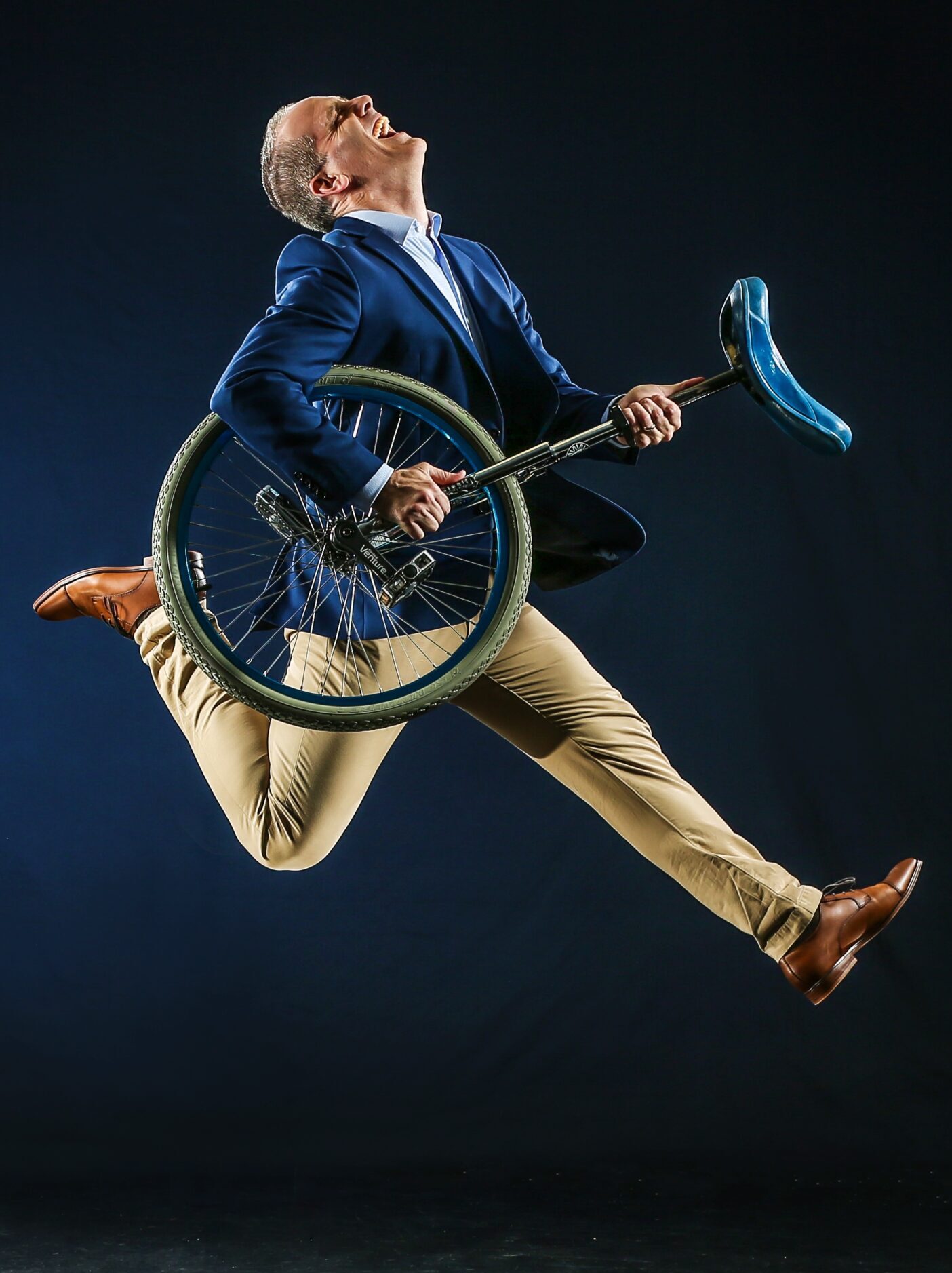 A man in a blue blazer and beige pants leaps mid-air while holding a unicycle. He wears brown shoes and glasses, and poses energetically against a dark studio background.