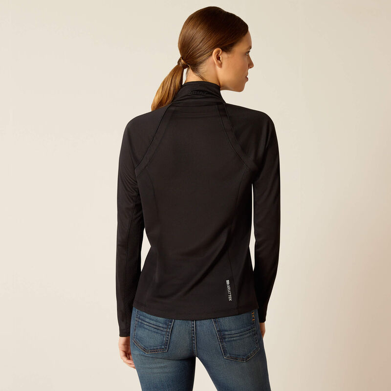 A woman with brown hair in a low ponytail is wearing a black long-sleeve zip-up jacket and blue jeans, standing with her back to the camera against a plain light background.