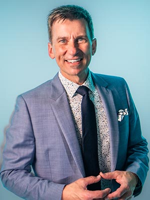 A smiling man wearing a blue suit, a patterned shirt, and a dark tie.