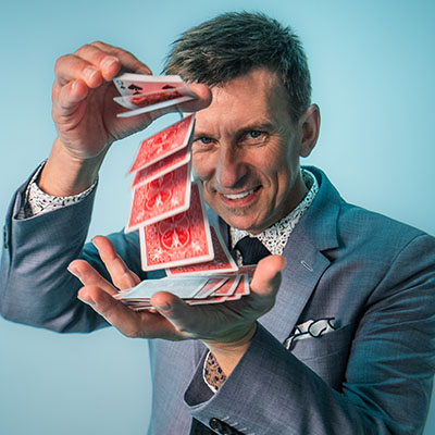 A magician in a suit smiles as he performs a waterfall card flourish with a deck of red playing cards.